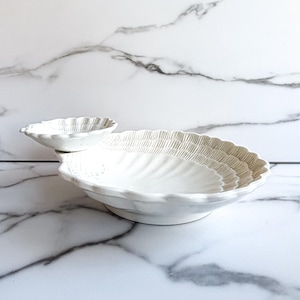 May include: A white ceramic serving dish shaped like a large scallop shell with a smaller, attached shell-shaped bowl on one side. The surface features textured, radiating ridges and a scalloped edge, finished with a glossy glaze.