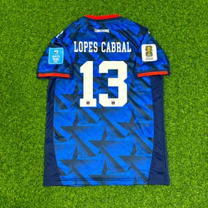 May include: A blue soccer jersey featuring a dark blue star pattern, the text LOPES CABRAL, and the number 13 in white. The shirt includes a CABO VERDE label, a FIFA World Cup 2026 patch, and a blue FIFA Football for Peace patch on the sleeves.