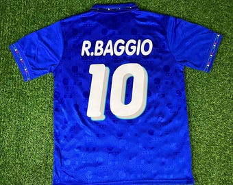 May include: A blue short-sleeved sports jersey featuring the name R.BAGGIO and the number 10 printed in white on the back. The fabric has a subtle tonal pattern, and the sleeve cuffs are accented with a small green, white, and red geometric trim.