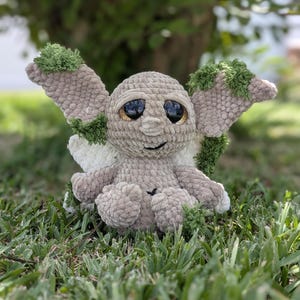 May include: A plush creature made of light beige yarn with large, dark, glossy eyes and a small stitched mouth. It features oversized, pointed ears accented with textured green yarn patches and small white wings on its back, crafted in a soft, fuzzy material.