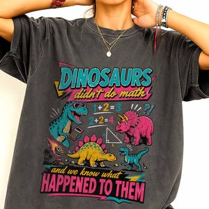 May include: A charcoal gray short-sleeve t-shirt featuring a colorful graphic with the text "DINOSAURS didn't do math" and "and we know what HAPPENED TO THEM". The design includes illustrations of three dinosaurs, math equations, and a meteor.