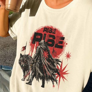 May include: A cream-coloured t-shirt featuring a graphic print of a hooded figure in a tattered cloak standing next to a wolf. The design includes a large red sun, red starburst accents, and the word "RISE" printed in bold black capital letters.