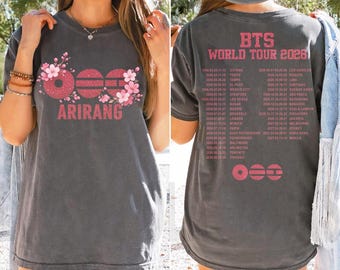 Op de afbeelding: Een antracietgrijs t-shirt met een vooropdruk van roze glittercirkels, kersenbloesems en de tekst ARIRANG. De achterkant toont BTS WORLD TOUR 2026 in roze letters met een lijst van tourdata en bijbehorende steden.