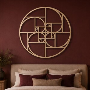May include: A large circular wooden wall art piece featuring a geometric golden ratio spiral design mounted on a dark red wall above a bed with beige bedding, dark red accent pillows, and a dark red throw blanket in a bedroom setting.