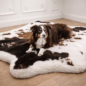 May include: A plush, faux fur area rug featuring an irregular, organic shape with a cowhide-inspired pattern in shades of white, dark brown, and tan. The rug has a thick, soft texture and is displayed with a medium-sized dog resting on its surface.
