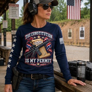 May include: A navy blue long-sleeve shirt featuring a graphic of a scroll with the text We The People, a rifle, and the text The Constitution Is My Permit. The sleeves have white stars and a flag patch. A black tumbler with text is also visible.