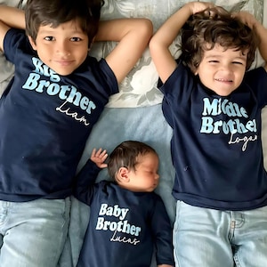 May include: Three brothers wearing navy blue shirts with the words "Big Brother", "Middle Brother", and "Baby Brother" in light blue. The shirts are personalized with the names Liam, Logan, and Lucas. The brothers are lying on a bed with a floral patterned sheet.