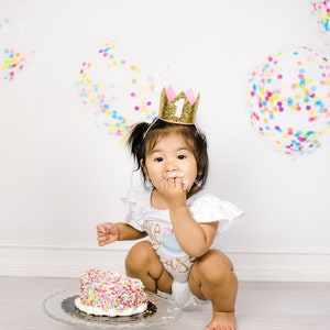 Pu&ograve; includere: Un bambino piccolo che indossa una corona dorata con glitter e un numero uno rosa, &egrave; seduto sul pavimento e mangia una torta con codette colorate. Il bambino indossa una tutina bianca con le parole "Cake Smash" su di essa. Lo sfondo &egrave; bianco con palloncini di coriandoli colorati.