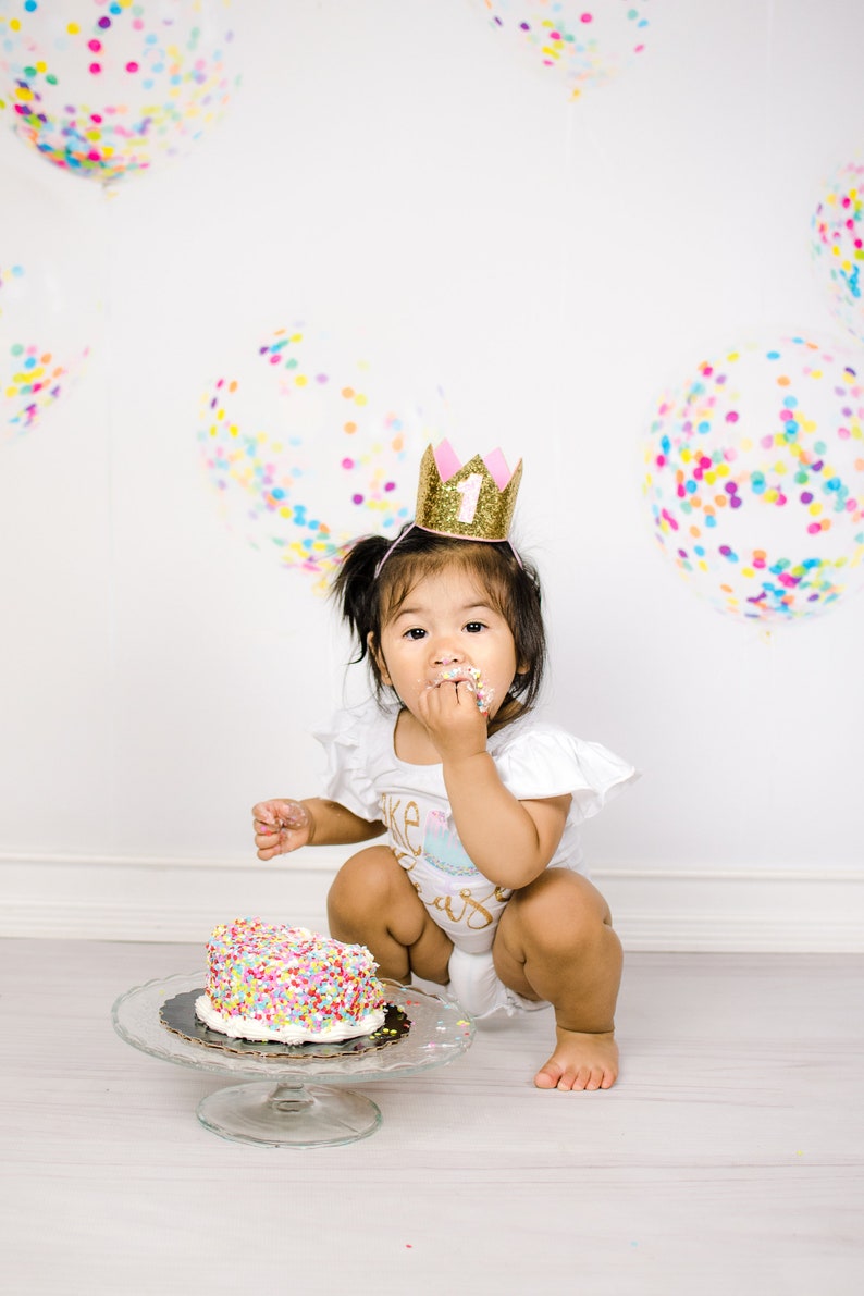 Pu&ograve; includere: Un bambino piccolo che indossa una corona dorata con glitter e un numero uno rosa, &egrave; seduto sul pavimento e mangia una torta con codette colorate. Il bambino indossa una tutina bianca con le parole "Cake Smash" su di essa. Lo sfondo &egrave; bianco con palloncini di coriandoli colorati.