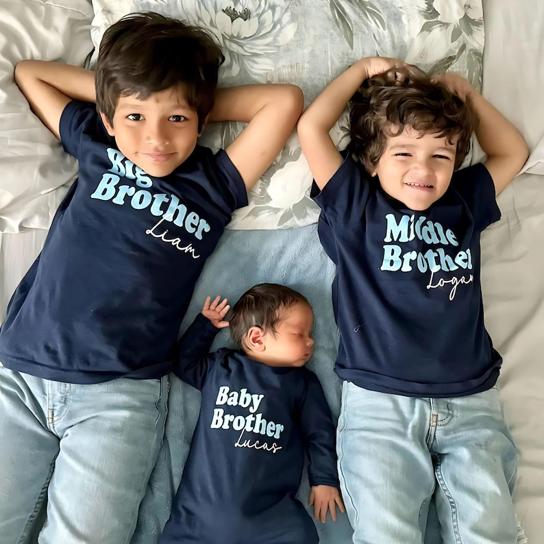 Op de afbeelding: Drie broers dragen donkerblauwe shirts met de woorden "Grote Broer", "Middelste Broer" en "Kleine Broer" in lichtblauw. De shirts zijn gepersonaliseerd met de namen Liam, Logan en Lucas. De broers liggen op een bed met een bloemenprint.