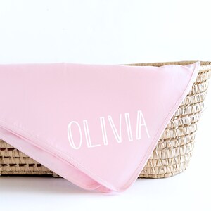 May include: A pink baby blanket with the name "Olivia" embroidered in white on it. The blanket is folded and placed in a wicker basket.