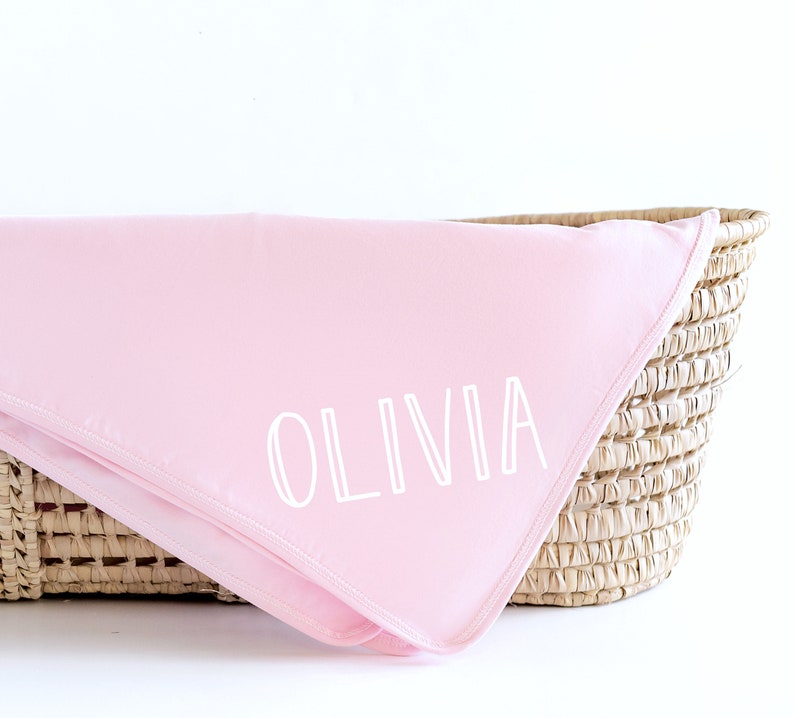 May include: A pink baby blanket with the name "Olivia" embroidered in white on it. The blanket is folded and placed in a wicker basket.