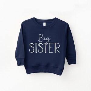 May include: A navy blue sweatshirt with the words "Big Sister" in white script and block letters. The sweatshirt has a crew neck and long sleeves. The text is centered on the front of the garment.