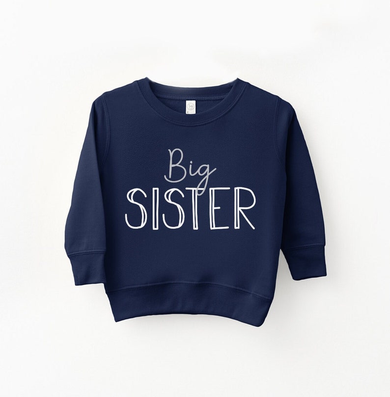 May include: A navy blue sweatshirt with the words "Big Sister" in white script and block letters. The sweatshirt has a crew neck and long sleeves. The text is centered on the front of the garment.