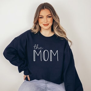 May include: Navy blue sweatshirt with the text "the MOM" in white. The word "the" is in a cursive font, and "MOM" is in a bold, block letter font. The sweatshirt is a crew neck style.