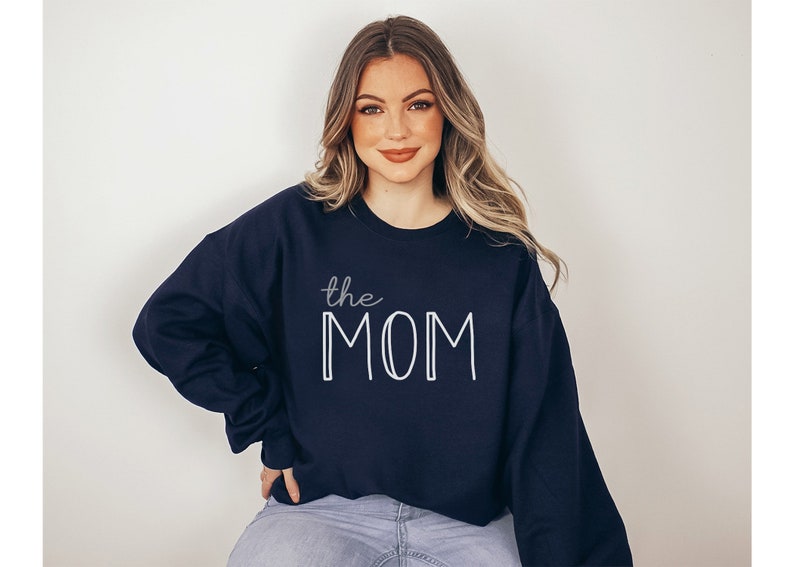 May include: Navy blue sweatshirt with the text "the MOM" in white. The word "the" is in a cursive font, and "MOM" is in a bold, block letter font. The sweatshirt is a crew neck style.