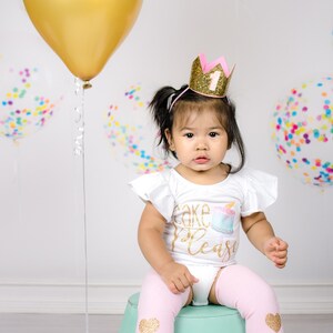 Pu&ograve; includere: Una bambina che indossa una maglietta bianca con la scritta "Cake Please" in glitter dorati e una corona di glitter rosa e oro con il numero "1" sopra. &Egrave; seduta su un secchio verde con leggings rosa con cuori di glitter dorati.