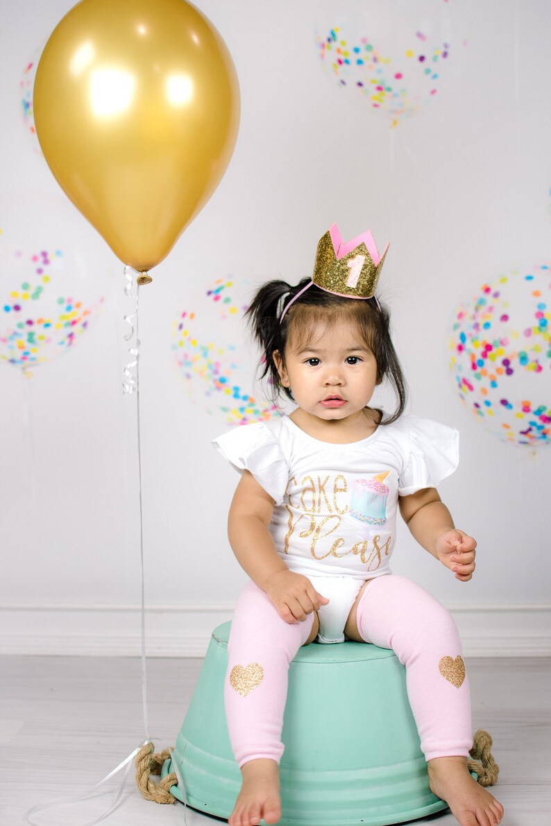 Pu&ograve; includere: Una bambina che indossa una maglietta bianca con la scritta "Cake Please" in glitter dorati e una corona di glitter rosa e oro con il numero "1" sopra. &Egrave; seduta su un secchio verde con leggings rosa con cuori di glitter dorati.