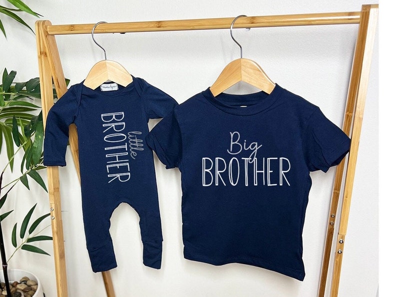 May include: Navy blue baby clothes set, including a long-sleeved onesie with "little brother" text and a short-sleeved t-shirt with "Big Brother" text. Both items are hanging on a wooden rack.