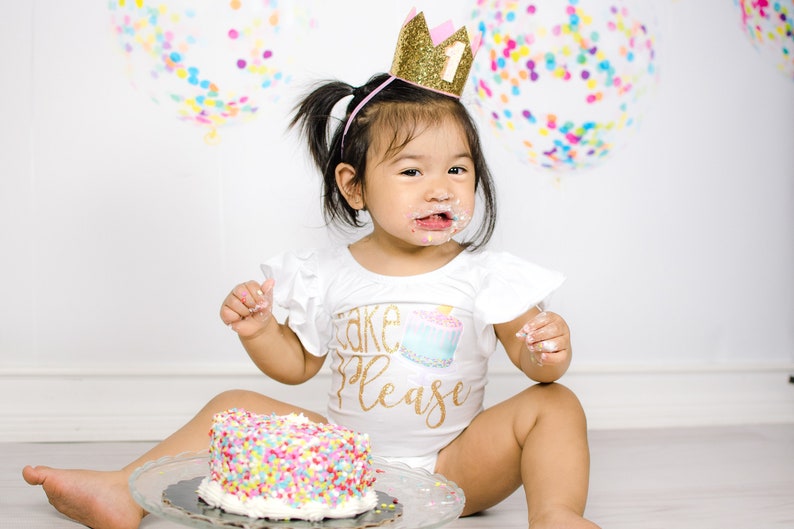 Pu&ograve; includere: Una bambina con una maglietta bianca con la scritta "Cake Please" e una corona dorata con glitter e un numero uno rosa in cima, &egrave; seduta sul pavimento con una torta bianca ricoperta di codette colorate.