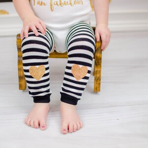 Puede incluir: Calentadores de piernas para beb&eacute; a rayas blancas y negras con aplicaciones de coraz&oacute;n con purpurina dorada. El beb&eacute; lleva los calentadores y est&aacute; sentado en una peque&ntilde;a silla de madera. El beb&eacute; lleva un body blanco con texto dorado "I am fabulous".