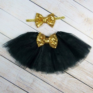 Puede incluir: Una falda tut&uacute; de tul negro con un gran lazo de lentejuelas doradas y una diadema a juego con un lazo de lentejuelas doradas. La falda y la diadema se muestran sobre una superficie de madera clara.