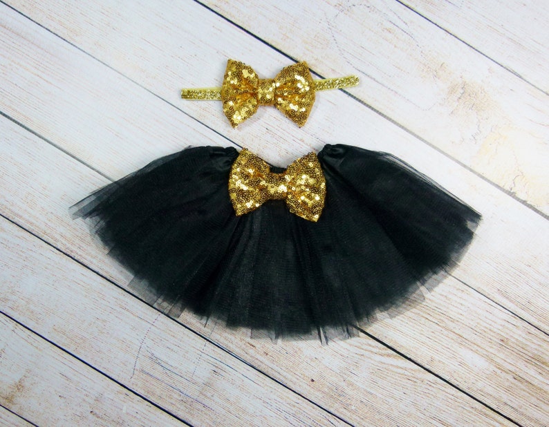 Puede incluir: Una falda tut&uacute; de tul negro con un gran lazo de lentejuelas doradas y una diadema a juego con un lazo de lentejuelas doradas. La falda y la diadema se muestran sobre una superficie de madera clara.
