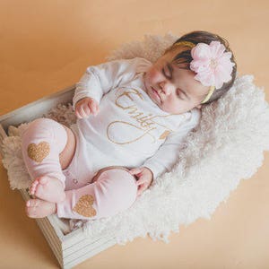 Puede incluir: Una ni&ntilde;a beb&eacute; con un body blanco con letras doradas brillantes que dicen "Emily" y calentadores de piernas rosas con corazones dorados brillantes. Lleva una diadema con una flor rosa y est&aacute; tumbada en una manta blanca y esponjosa en una caja de madera.