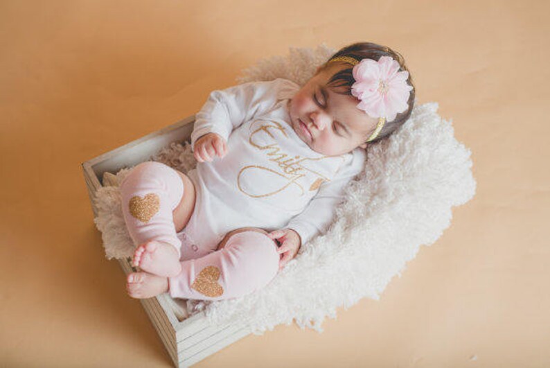 Puede incluir: Una ni&ntilde;a beb&eacute; con un body blanco con letras doradas brillantes que dicen "Emily" y calentadores de piernas rosas con corazones dorados brillantes. Lleva una diadema con una flor rosa y est&aacute; tumbada en una manta blanca y esponjosa en una caja de madera.