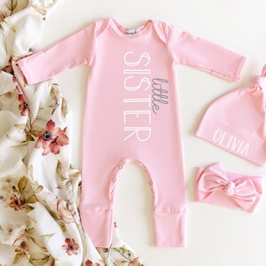May include: Pink baby outfit set with a long-sleeved onesie, a hat, and a headband. The onesie has the text "little sister" printed on it. The hat has the name "Olivia" printed on it.