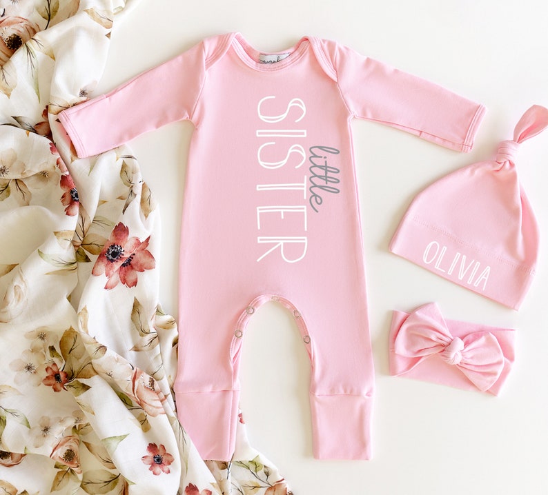 May include: Pink baby outfit set with a long-sleeved onesie, a hat, and a headband. The onesie has the text "little sister" printed on it. The hat has the name "Olivia" printed on it.