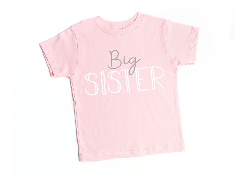 May include: Pink short-sleeved t-shirt with the text "Big Sister" in white and gray lettering.