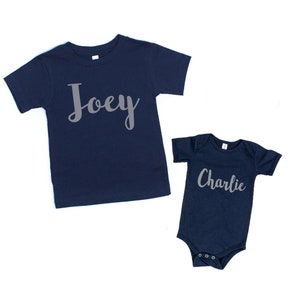 Może przedstawiać: Dwie koszulki w kolorze granatowym z nadrukiem imion "Joey" i "Charlie" w kolorze szarym z przodu.