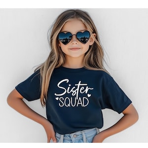 May include: A navy blue t-shirt with the words "Sister Squad" in white script. The shirt is worn by a young person wearing heart-shaped sunglasses. The text is in a playful font, with small hearts accenting the word "Squad".