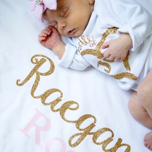 Puede incluir: Una manta de beb&eacute; blanca con el nombre "Reagan Rose" escrito en letras doradas con purpurina. Un beb&eacute; est&aacute; durmiendo en la manta con un mono blanco con letras doradas con purpurina y un gorro a rayas rosa y blanco con un strass.