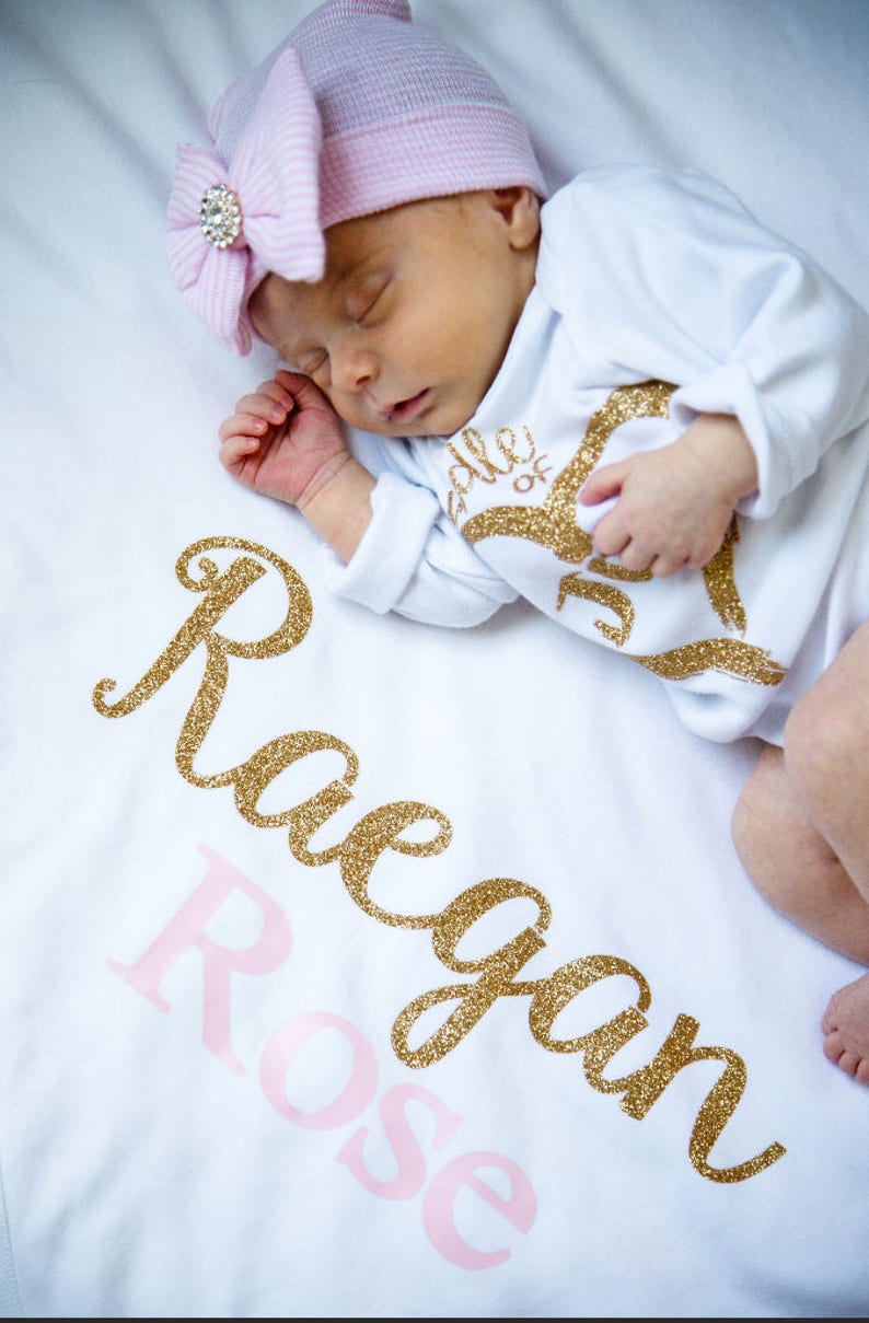 Puede incluir: Una manta de beb&eacute; blanca con el nombre "Reagan Rose" escrito en letras doradas con purpurina. Un beb&eacute; est&aacute; durmiendo en la manta con un mono blanco con letras doradas con purpurina y un gorro a rayas rosa y blanco con un strass.