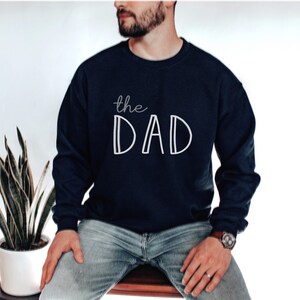 May include: Navy blue sweatshirt with the white text "the DAD". The sweatshirt is a long-sleeved crew neck style. The image shows a person wearing the sweatshirt.