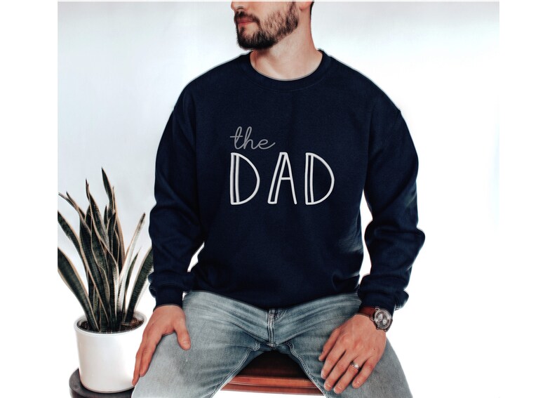 May include: Navy blue sweatshirt with the white text "the DAD". The sweatshirt is a long-sleeved crew neck style. The image shows a person wearing the sweatshirt.
