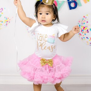 Pu&ograve; includere: Una bambina che indossa una maglietta bianca con la scritta "cake please" e una gonna di tulle rosa. La bambina indossa una corona dorata con la parte superiore rosa e il numero uno sopra. La bambina tiene uno spago e guarda la fotocamera.