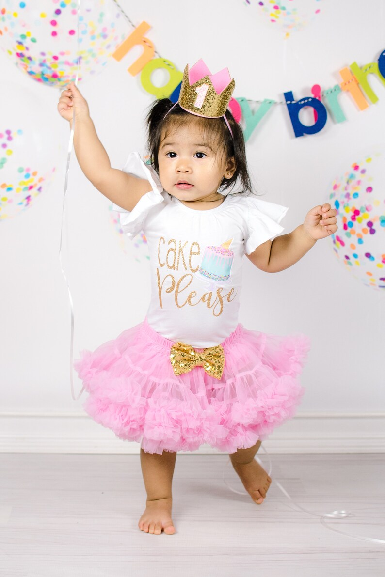 Pu&ograve; includere: Una bambina che indossa una maglietta bianca con la scritta "cake please" e una gonna di tulle rosa. La bambina indossa una corona dorata con la parte superiore rosa e il numero uno sopra. La bambina tiene uno spago e guarda la fotocamera.