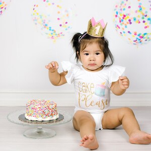 Pu&ograve; includere: Una bambina con una maglietta bianca con la scritta "Cake Please" e una corona dorata con il numero 1, seduta su un pavimento bianco con una torta decorata con codette colorate davanti a lei.
