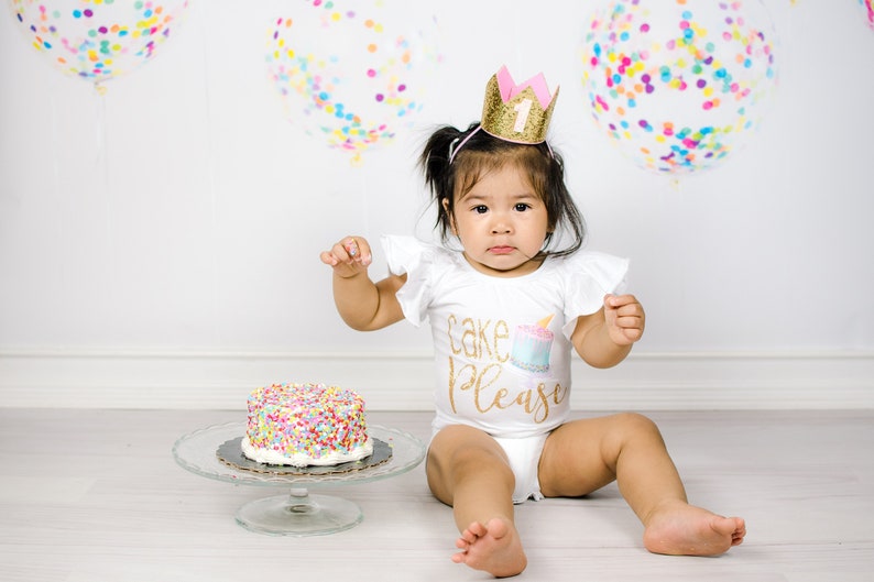 Pu&ograve; includere: Una bambina con una maglietta bianca con la scritta "Cake Please" e una corona dorata con il numero 1, seduta su un pavimento bianco con una torta decorata con codette colorate davanti a lei.