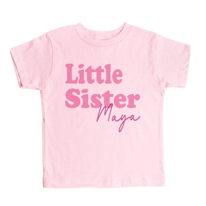 May include: Pink short-sleeved t-shirt with the text "Little Sister Maya" in pink lettering.