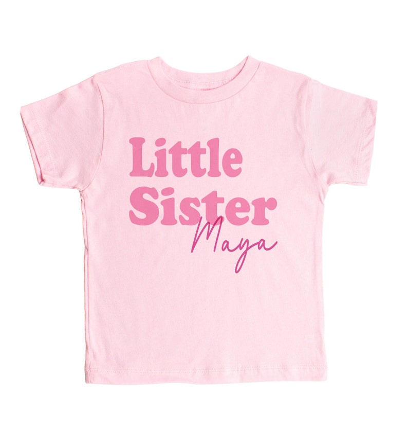 May include: Pink short-sleeved t-shirt with the text "Little Sister Maya" in pink lettering.