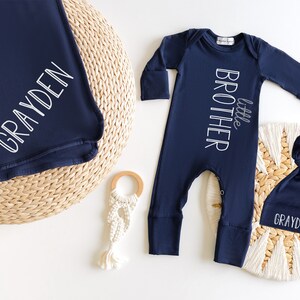 May include: Navy blue baby outfit set including a blanket, a long-sleeved romper with "little brother" text, and a knotted hat. The blanket and hat are personalized with the name "GRAYDEN" in white script. A wooden teether and macrame toy are also present.