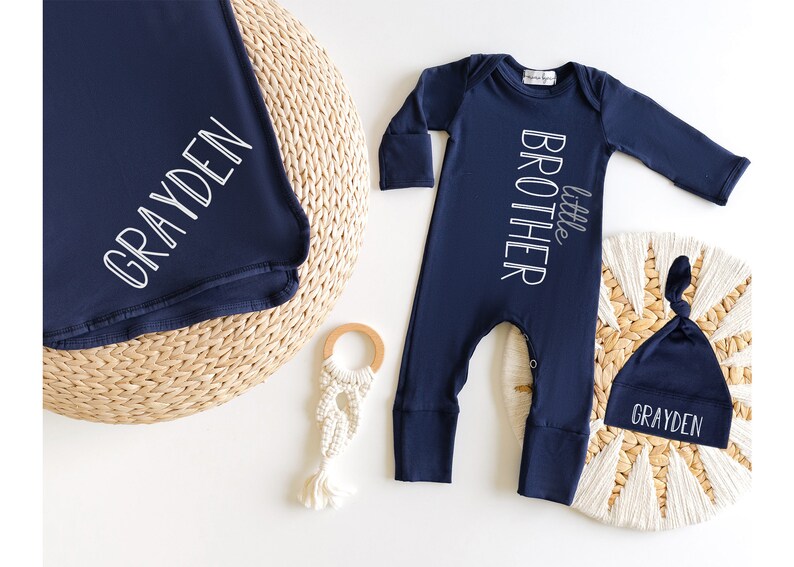 May include: Navy blue baby outfit set including a blanket, a long-sleeved romper with "little brother" text, and a knotted hat. The blanket and hat are personalized with the name "GRAYDEN" in white script. A wooden teether and macrame toy are also present.