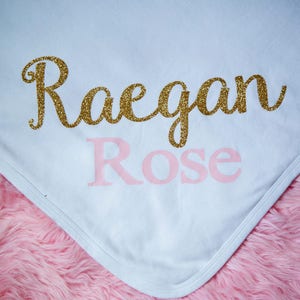 Puede incluir: Manta de beb&eacute; blanca con el nombre "Raegan Rose" escrito en purpurina dorada y letras rosas.