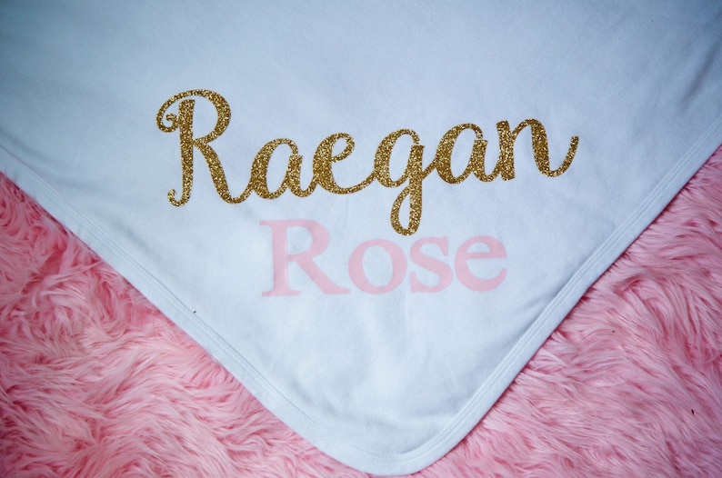 Puede incluir: Manta de beb&eacute; blanca con el nombre "Raegan Rose" escrito en purpurina dorada y letras rosas.