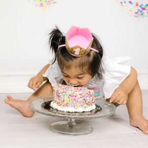 Pu&ograve; includere: Una bambina con una corona rosa con un '1' dorato, &egrave; seduta sul pavimento e mangia una torta ricoperta di codette colorate.