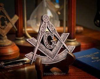 May include: A silver-toned metal Masonic square and compass with intricate details and a decorative stand. The compass features a letter "G" in the center.